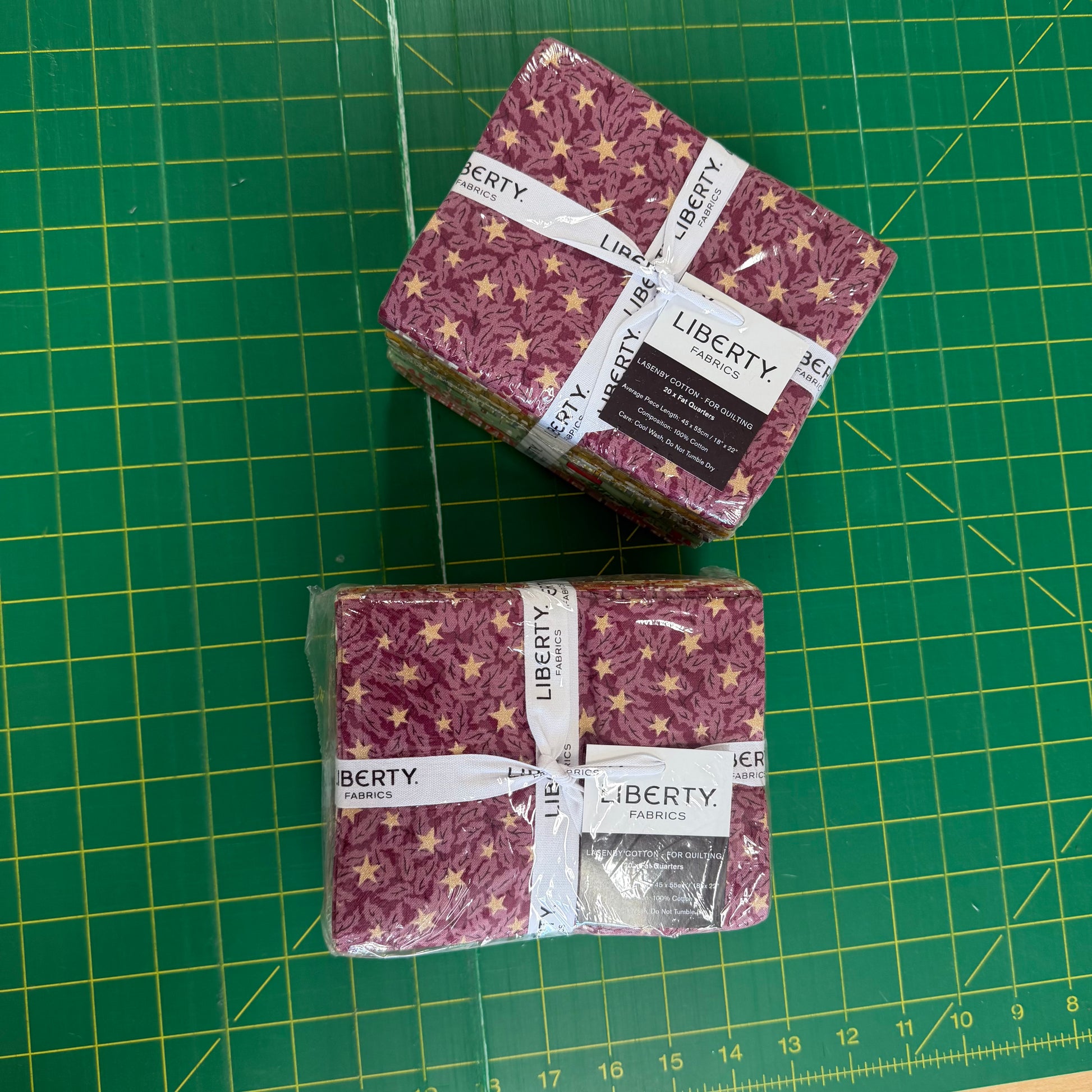 Two packaged stacks of Liberty Fabrics patterned fabric squares on a green cutting mat. The fabric has a dusty mauve/pink background with a gold star pattern.