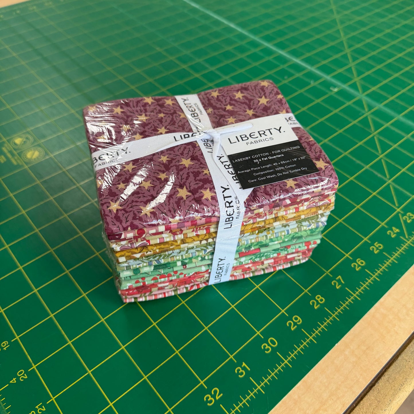A stack of Liberty Quilt Co. fabric bundles wrapped in clear plastic and tied with a white paper band, placed on a green cutting mat with measurement markings.