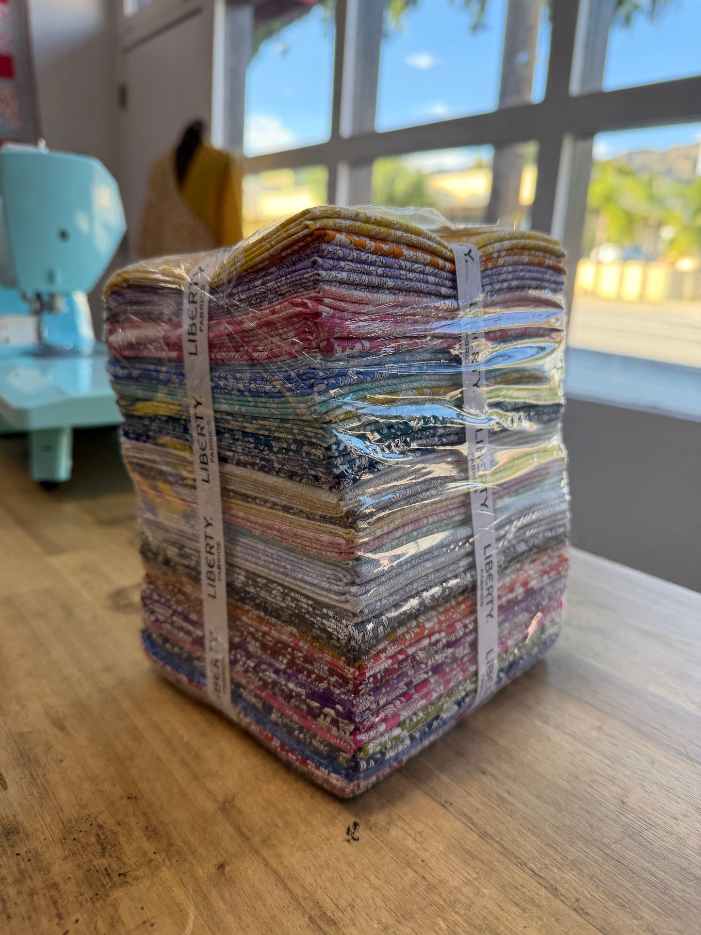 A stack of colorful fabric bundles wrapped in clear plastic and secured with white paper bands, sitting on a wooden table in a bright room with a sewing machine and window in the background.