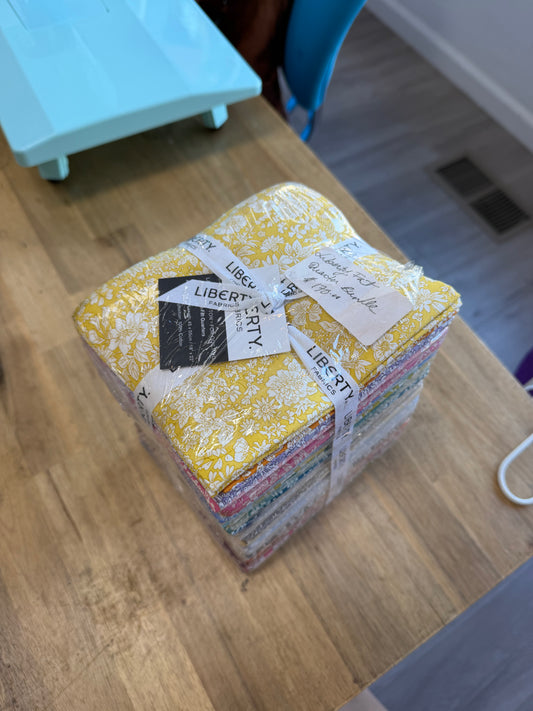 A stack of Liberty brand quilting fabric bundles wrapped in clear plastic and ribbon, resting on a wooden table.