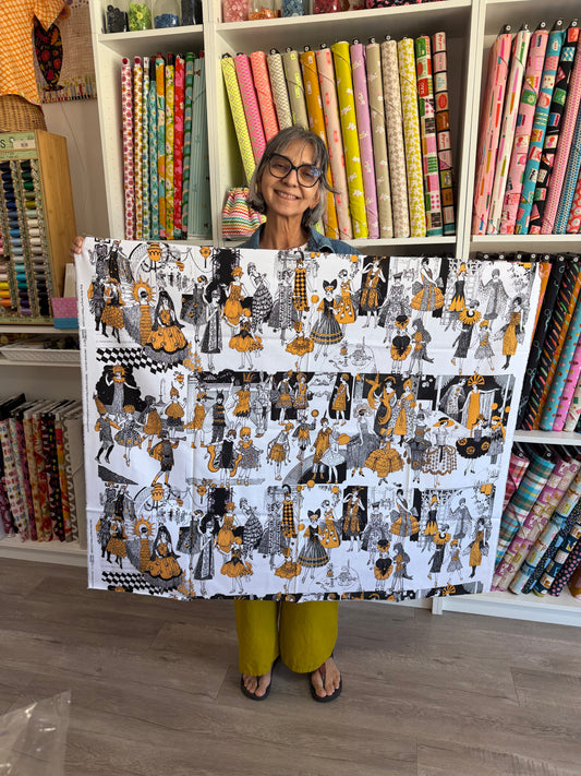 A woman holding up a large fabric panel featuring a whimsical black, white, and mustard yellow pattern of dancing figures, set against a background of shelves filled with colorful fabric rolls.