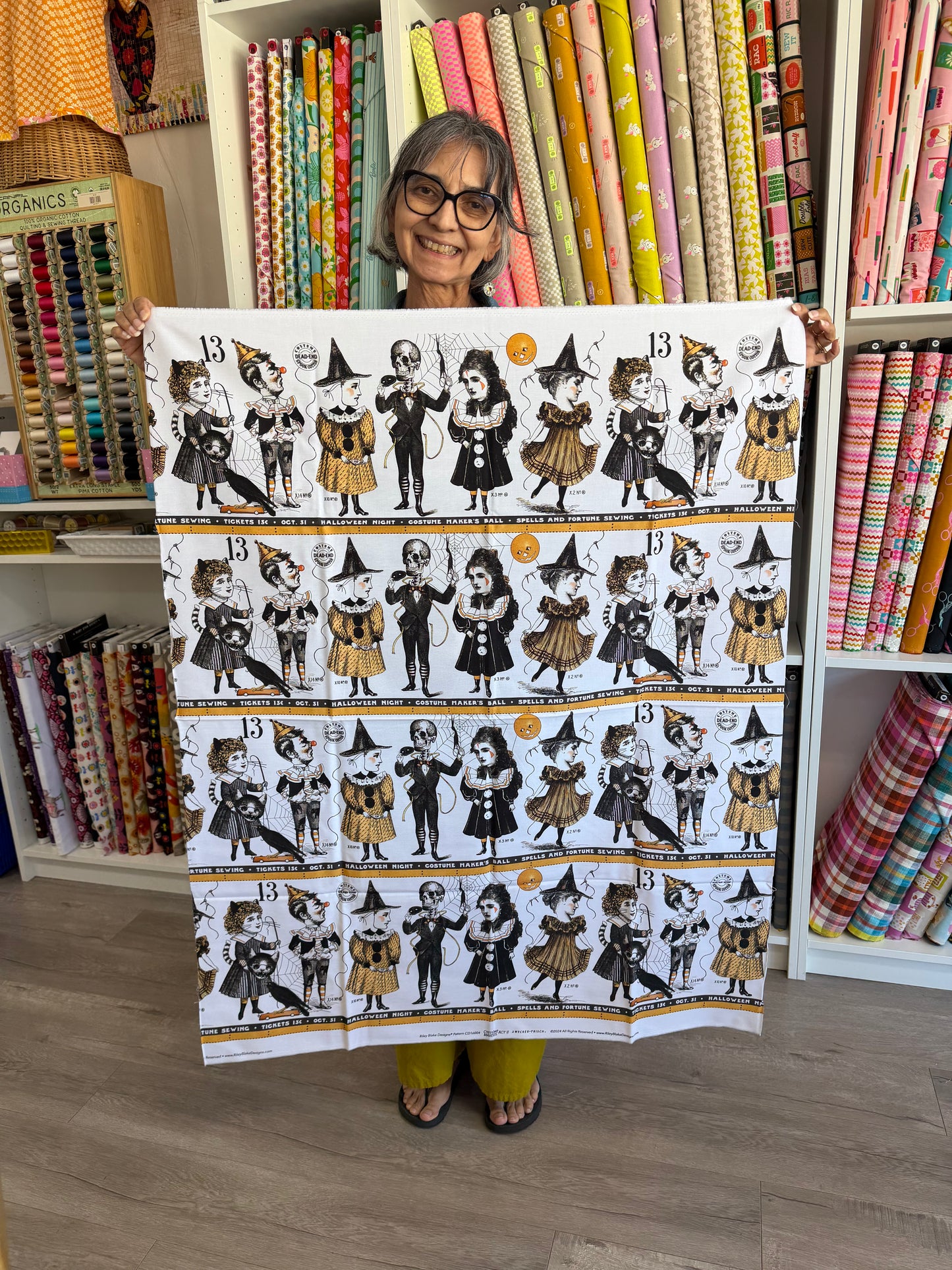 A woman holding up a large fabric panel featuring a repeating pattern of cartoon witch characters in black, white, and orange. The background shows shelves filled with colorful rolls of fabric.
