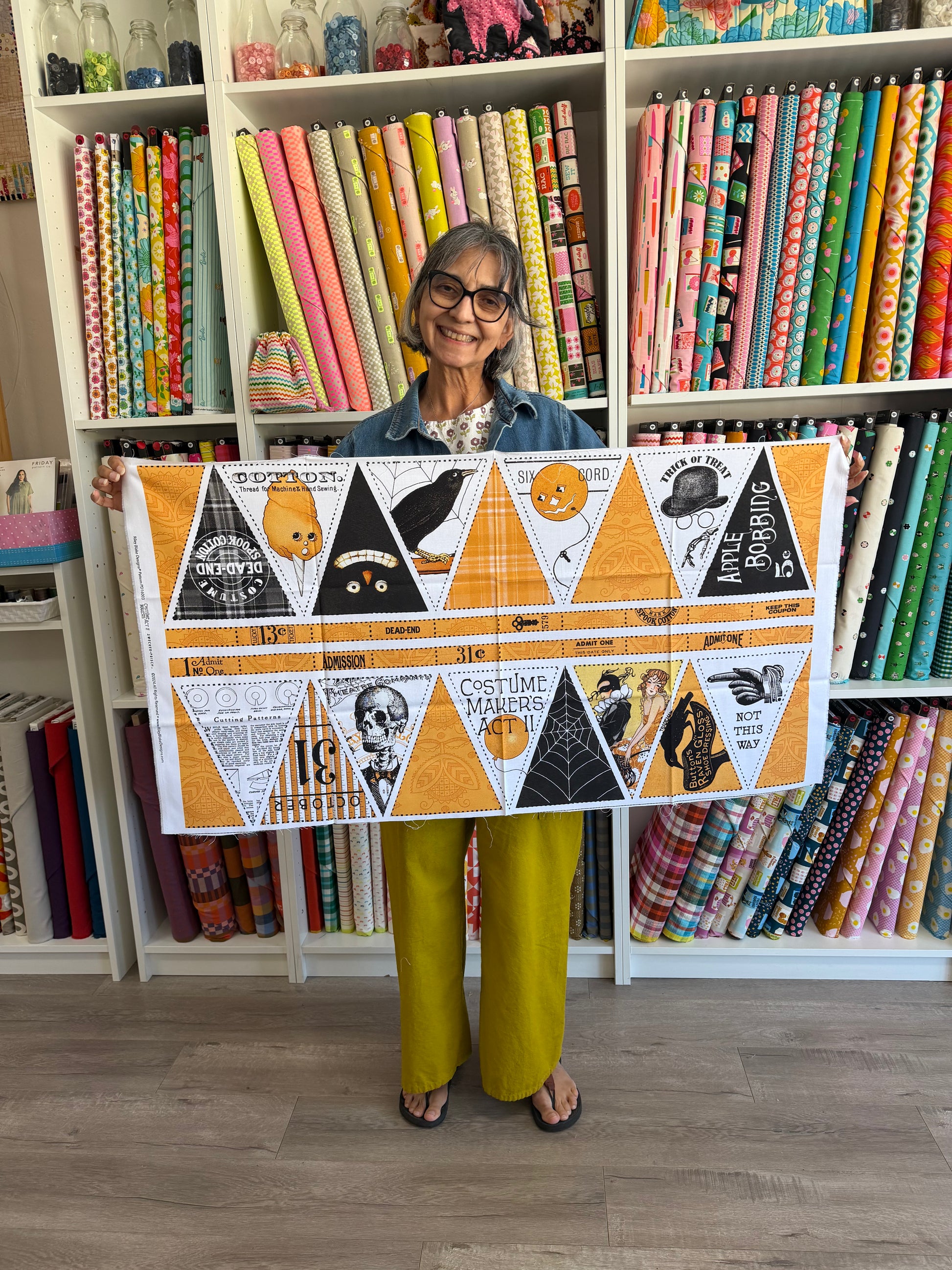 A woman holding up a large fabric banner with a Halloween-themed pattern, featuring orange, black, white, and yellow triangles with spiderwebs, ghosts, and spooky illustrations. She is standing in front of shelves filled with colorful fabric rolls.