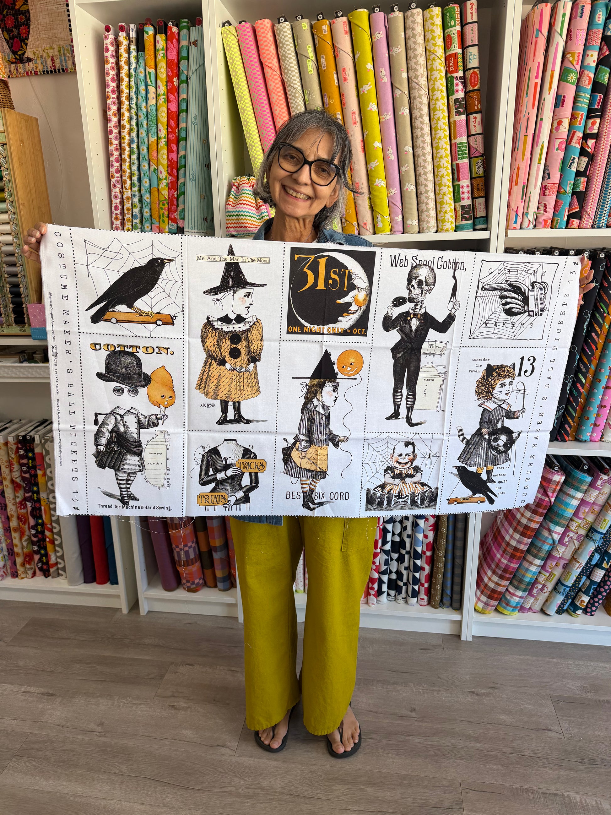 A woman wearing glasses and mustard yellow pants holds up a large white fabric print featuring black-and-white whimsical illustrations of crows, a witch, a cat, and vintage-style text. The background shows shelves filled with colorful rolls of fabric.