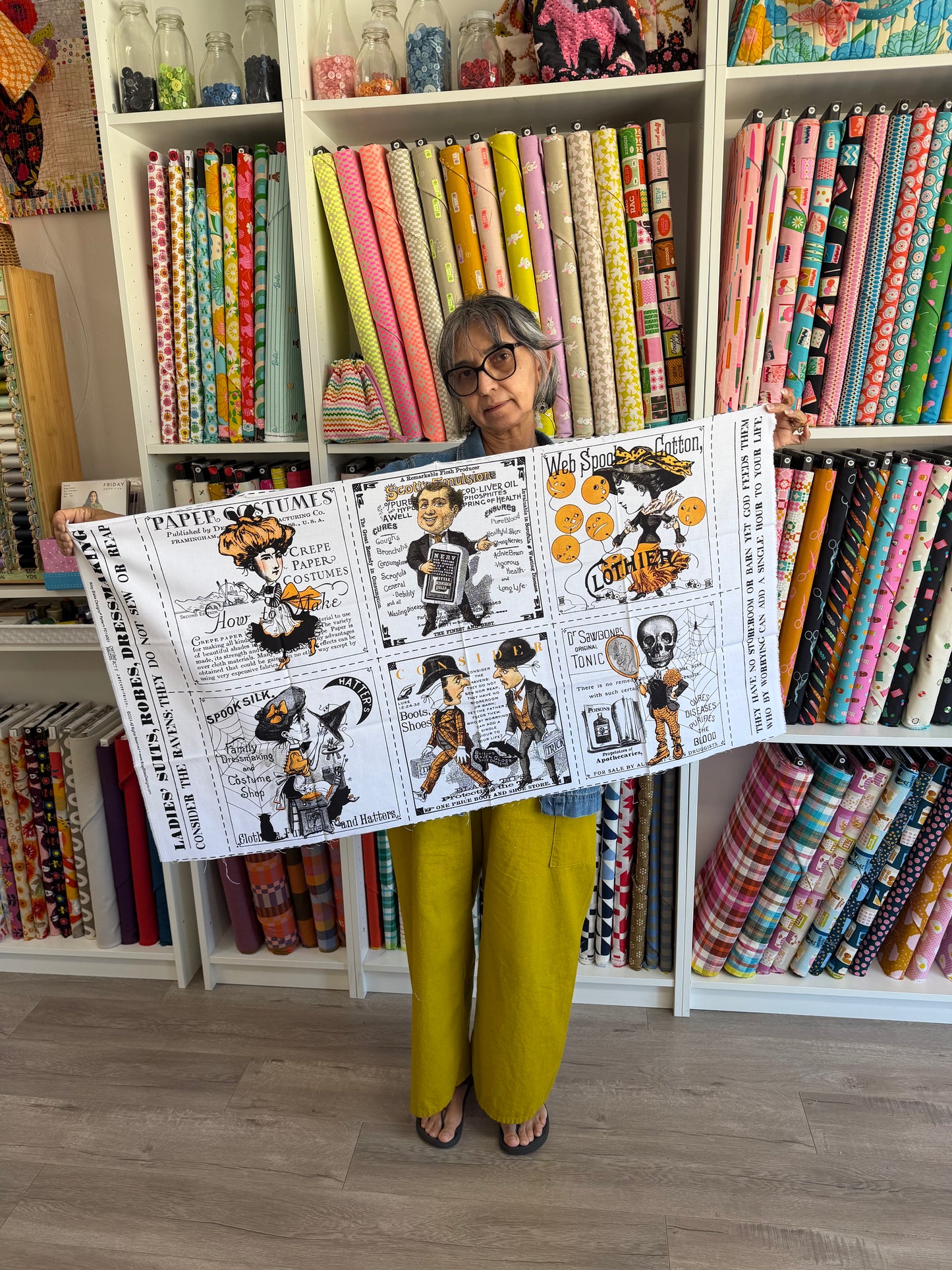 A woman wearing glasses and mustard yellow pants holds up a large white fabric banner featuring a black-and-white comic strip with colorful cartoon characters and text. The background shows shelves filled with colorful rolls of fabric in a craft or sewing supply store.