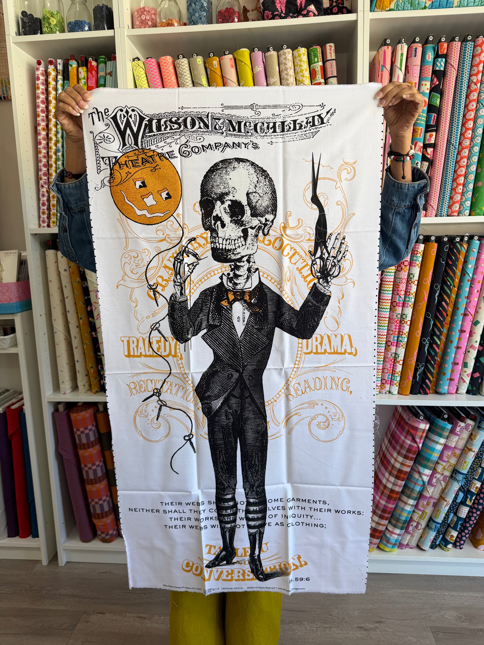 A person holds up a white fabric print featuring a vintage-style illustrated skeleton in a black suit with gold lettering. The background shows shelves filled with colorful rolls of fabric.