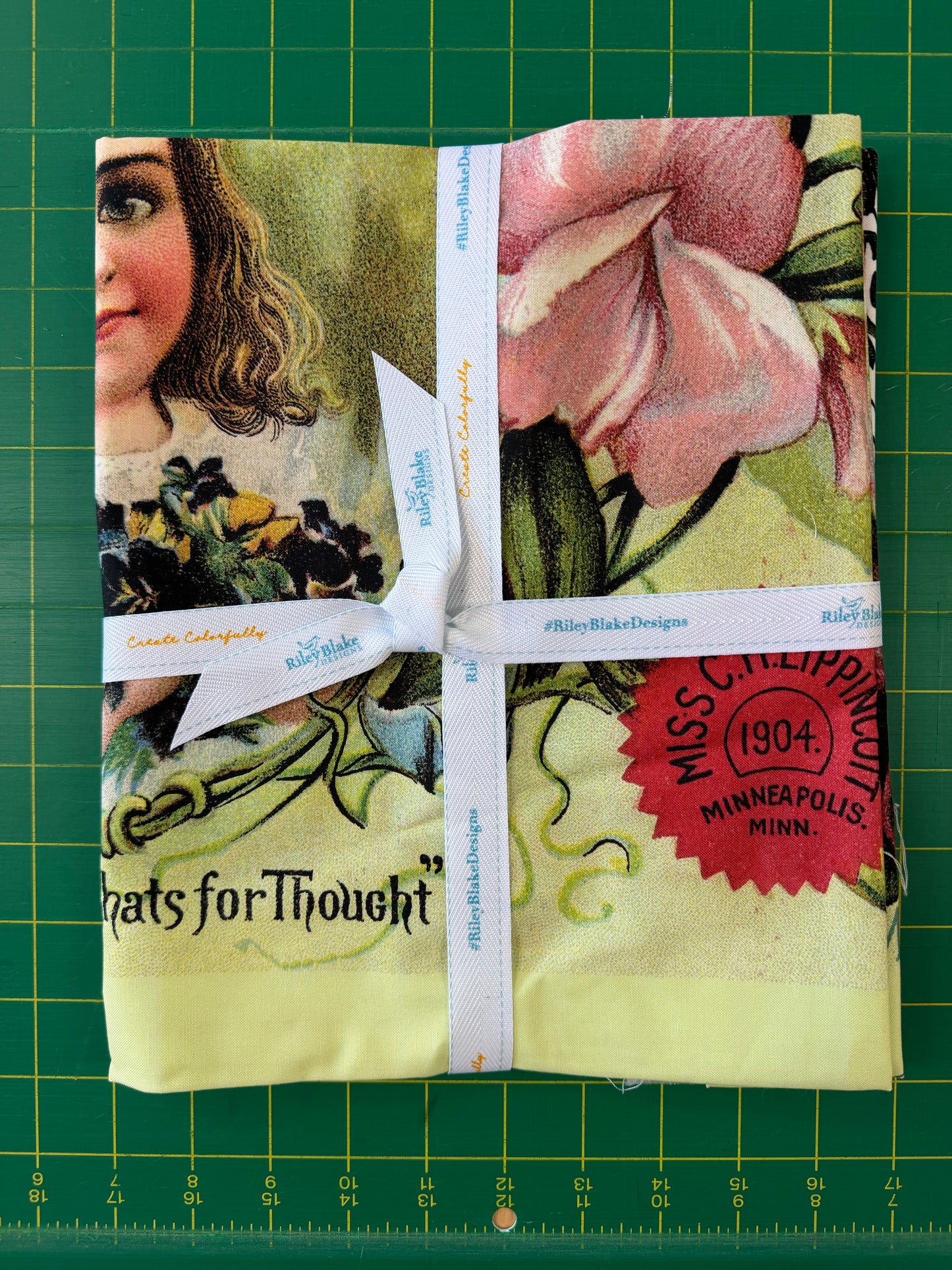 A folded vintage-style fabric bundle wrapped with a white ribbon, placed on a green cutting mat. The fabric features a collage print with a woman's face, a pink flower, black-and-white chain links, and a red circular stamp reading 'Miss C.