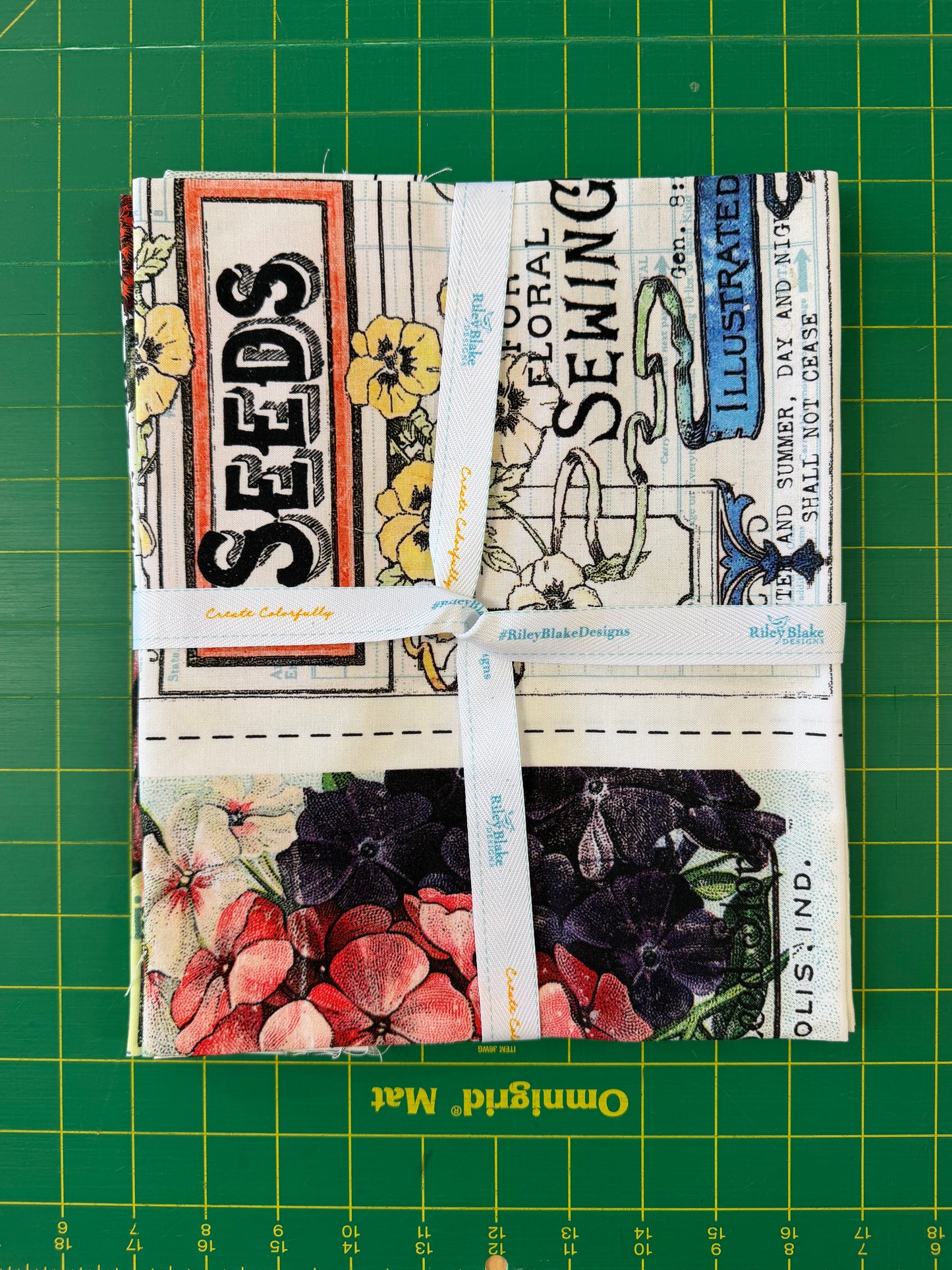 A folded fabric bundle with a vintage floral and botanical print, wrapped with a white ribbon, placed on a green cutting mat. The print features text like 'SEEDS' and 'SEWING' alongside yellow flowers and dark purple blossoms.