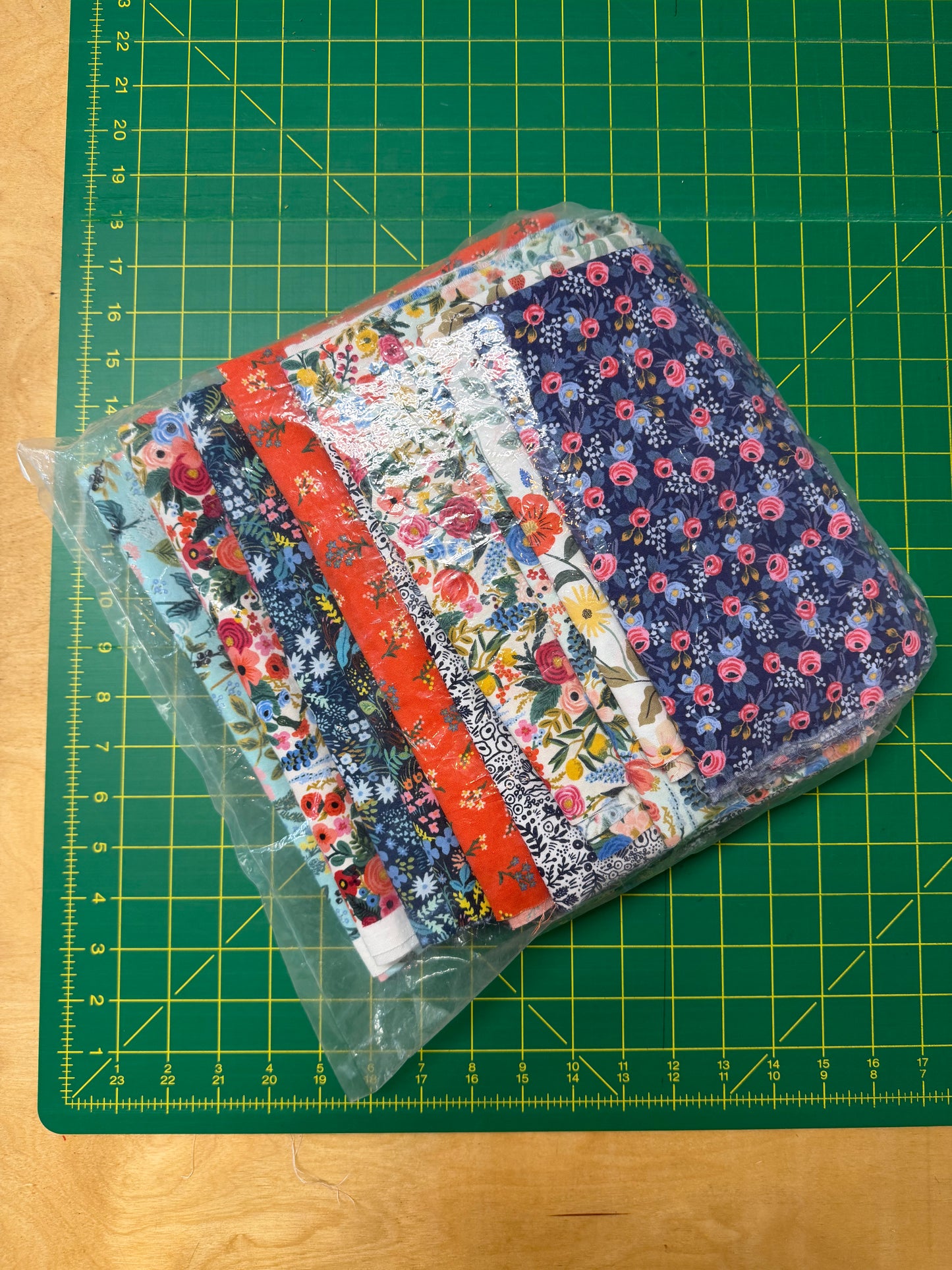A packaged set of floral fabric pouches or small fabric goods in clear plastic, placed on a green cutting mat over a wooden surface.