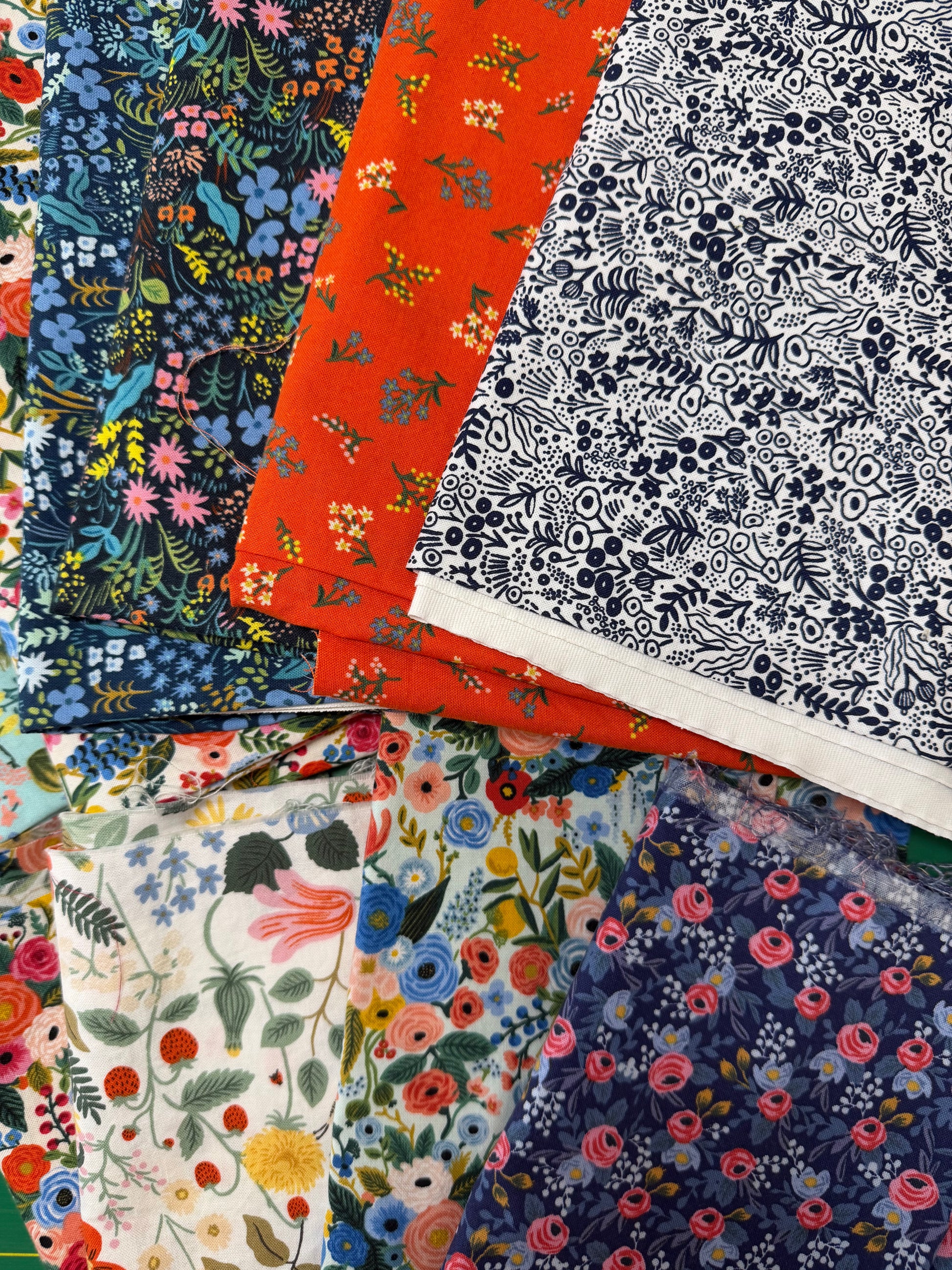 A stack of folded patterned fabric pieces displayed together, showing multiple floral and botanical prints in blue, orange, white, pink, yellow, and green.