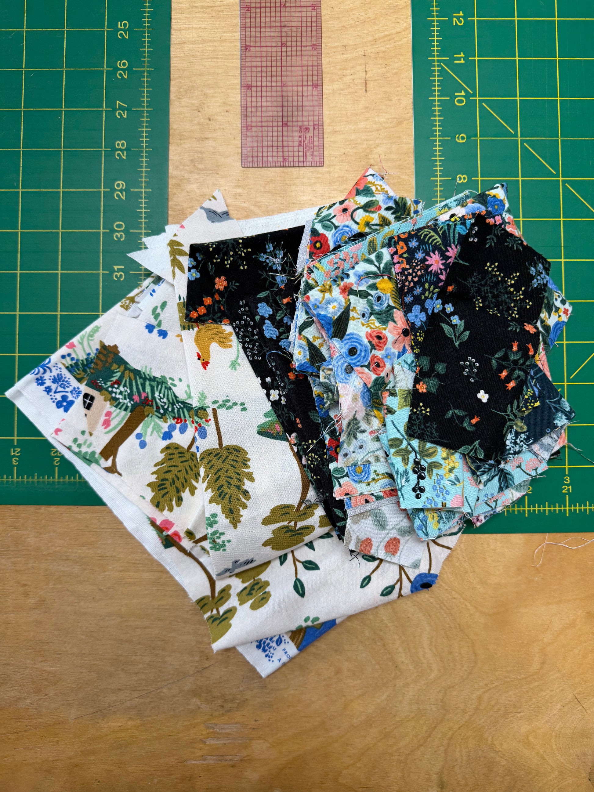 A pile of patterned fabric pieces laid on a wooden surface, with a green cutting mat and clear ruler visible in the background. The fabrics show various prints including floral, woodland, and abstract designs in black, white, blue, green, and pink.
