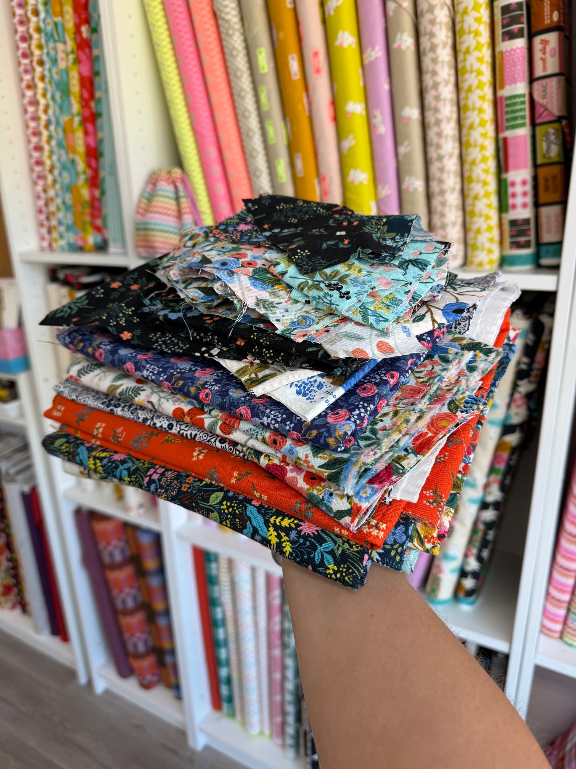 A hand holding a stack of colorful patterned fabric squares in front of shelves filled with rolls of fabric.