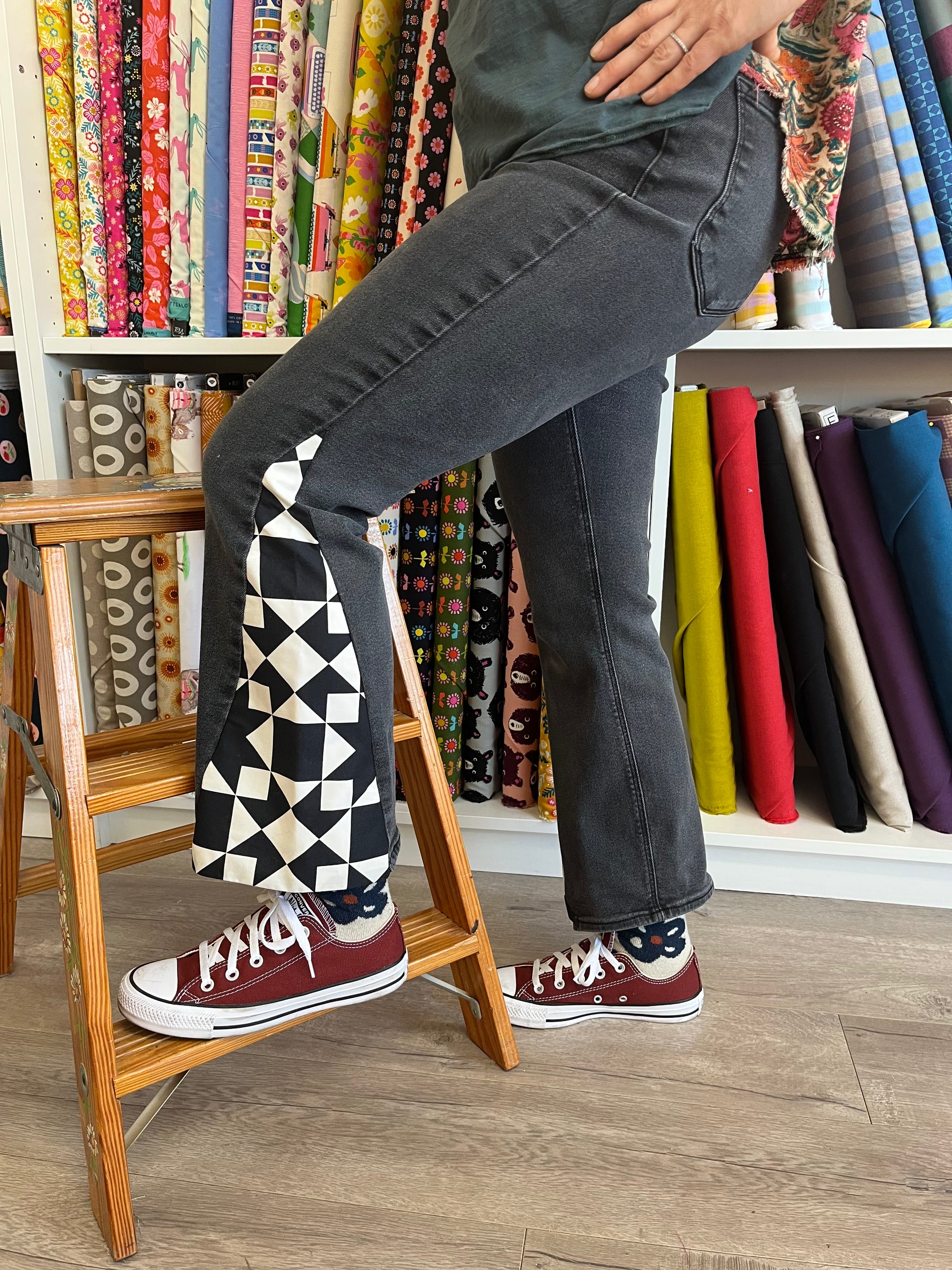 Person wearing dark jeans and maroon sneakers with a patterned sock, standing on a wooden ladder in front of fabric rolls.