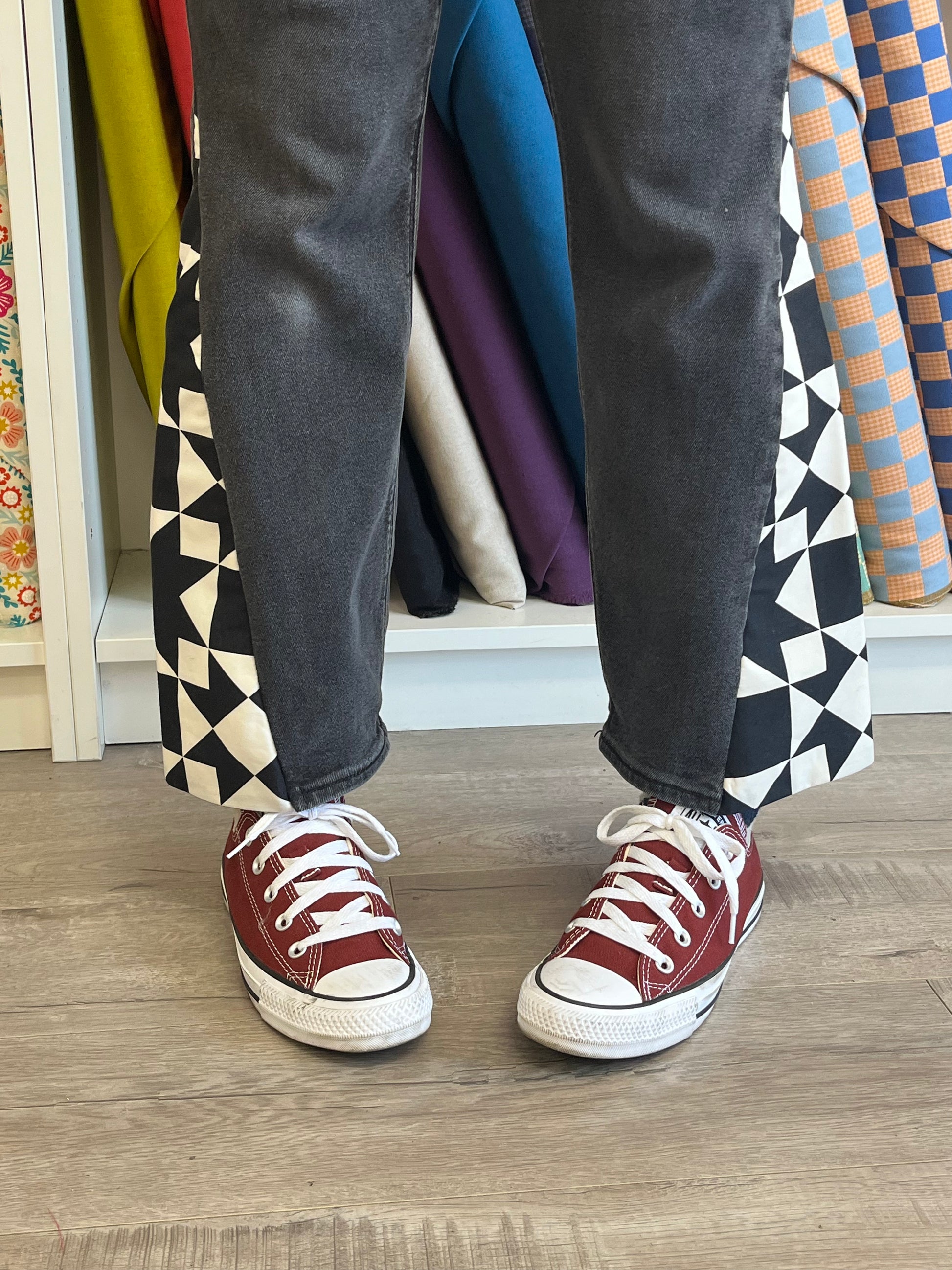 Person wearing maroon sneakers with white laces on a wooden floor, with colorful fabric rolls in the background.