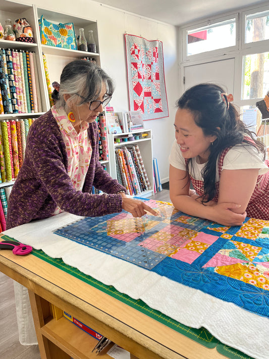 Two women collaborating in a bright craft room, working on a colorful patchwork quilt laid out on a wooden table. One woman points at the quilt while the other observes closely.