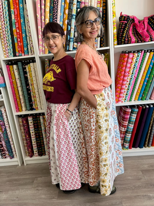 Two women wearing block printed skirts with shelves of colorful fabric behind them.