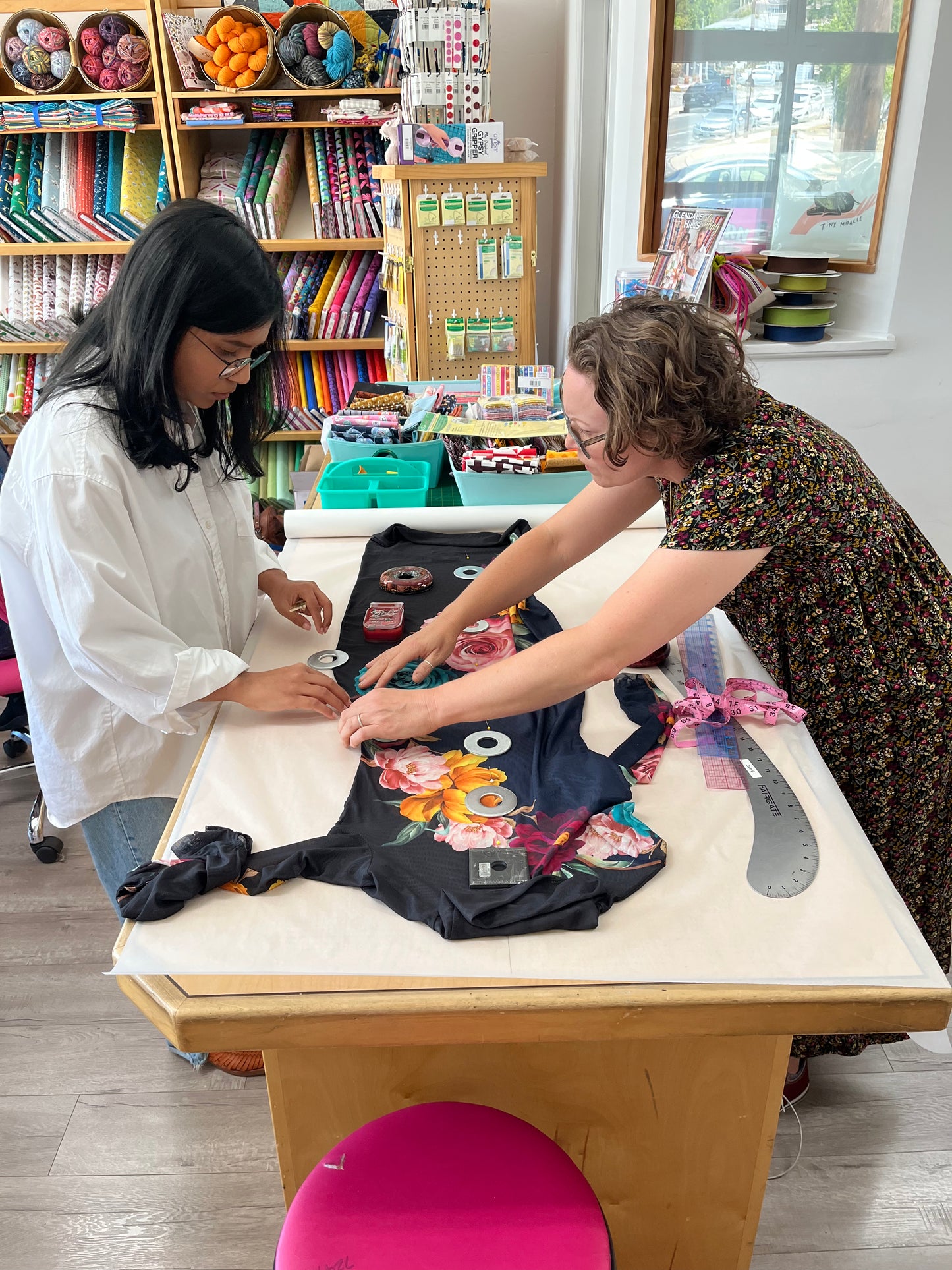 Two women working together at a table in a craft studio, assembling a black garment decorated with colorful floral patches and ribbon.