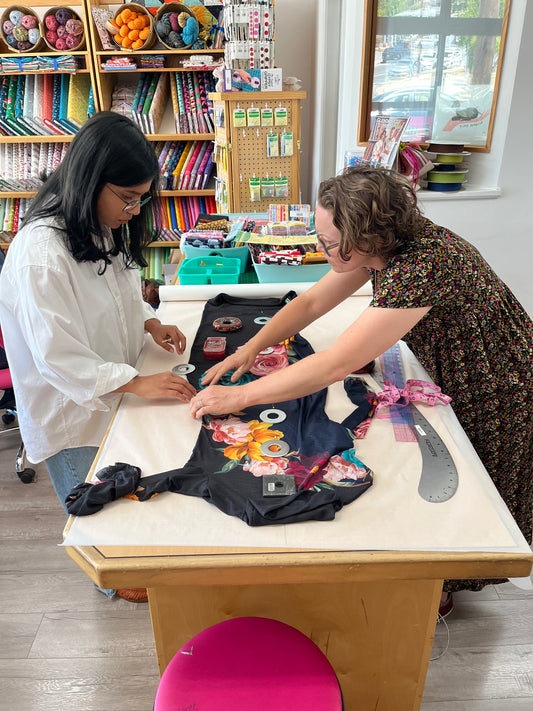Two women working together at a table in a craft studio, assembling a black garment decorated with colorful floral patches and ribbon.