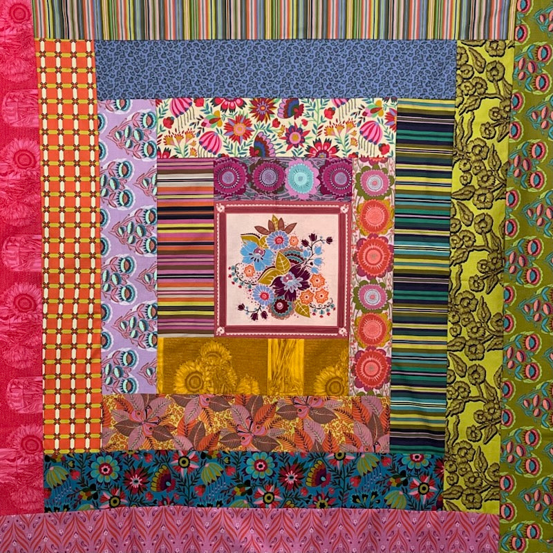 A colorful patchwork quilt or fabric artwork made of multiple patterned fabric panels arranged in concentric squares, including floral, striped, geometric, and paisley designs in pink, blue, green, orange, yellow, and purple.