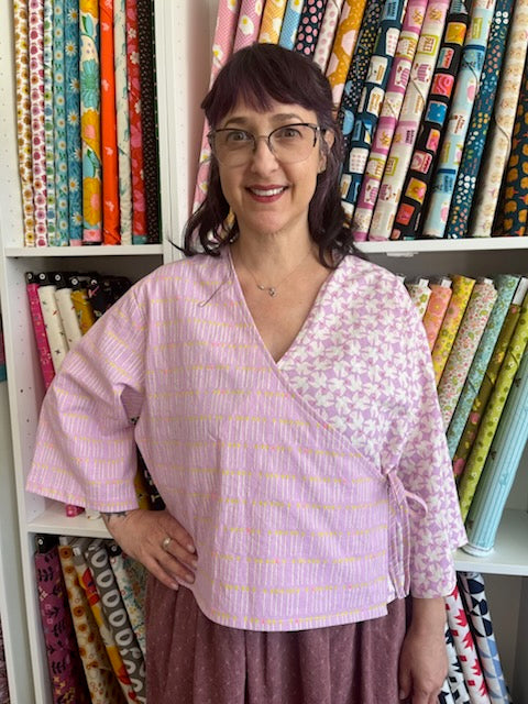A woman wearing a lavender wrap-style blouse with a textured vertical stripe pattern and a floral yoke, standing in front of shelves filled with colorful patterned fabric rolls.