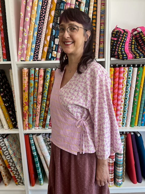 A smiling woman with glasses stands in front of shelves filled with colorful patterned fabric rolls, wearing a lavender patterned wrap-style top and a dark mauve skirt.
