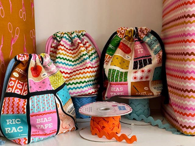 Three handmade drawstring bags made from colorful patchwork fabric with sewing-themed prints, displayed on a shelf with a spool of orange ric rac in front of them.