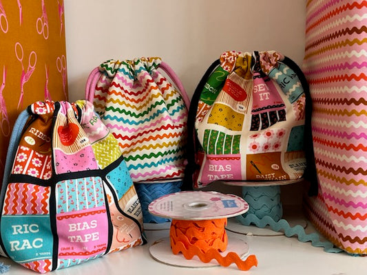 Three handmade drawstring bags made from colorful patchwork fabric with sewing-themed prints, displayed on a shelf with a spool of orange ric rac in front of them.