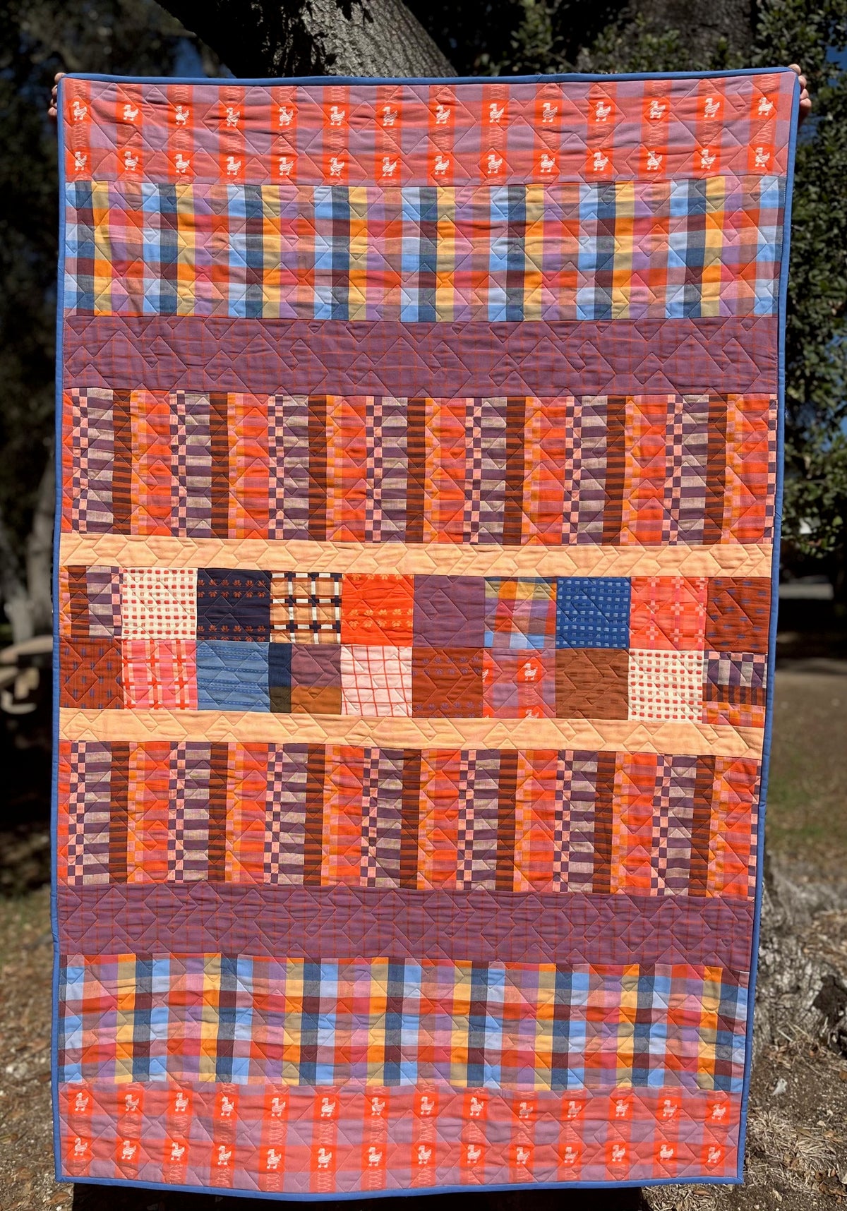 A colorful patchwork quilt with horizontal bands of orange, blue, brown, and beige patterns, displayed outdoors against a tree trunk and foliage.