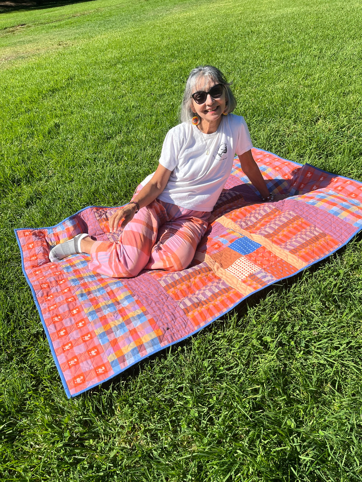 A woman wearing sunglasses sits cross-legged on a colorful patchwork quilt laid out on a green grass lawn.