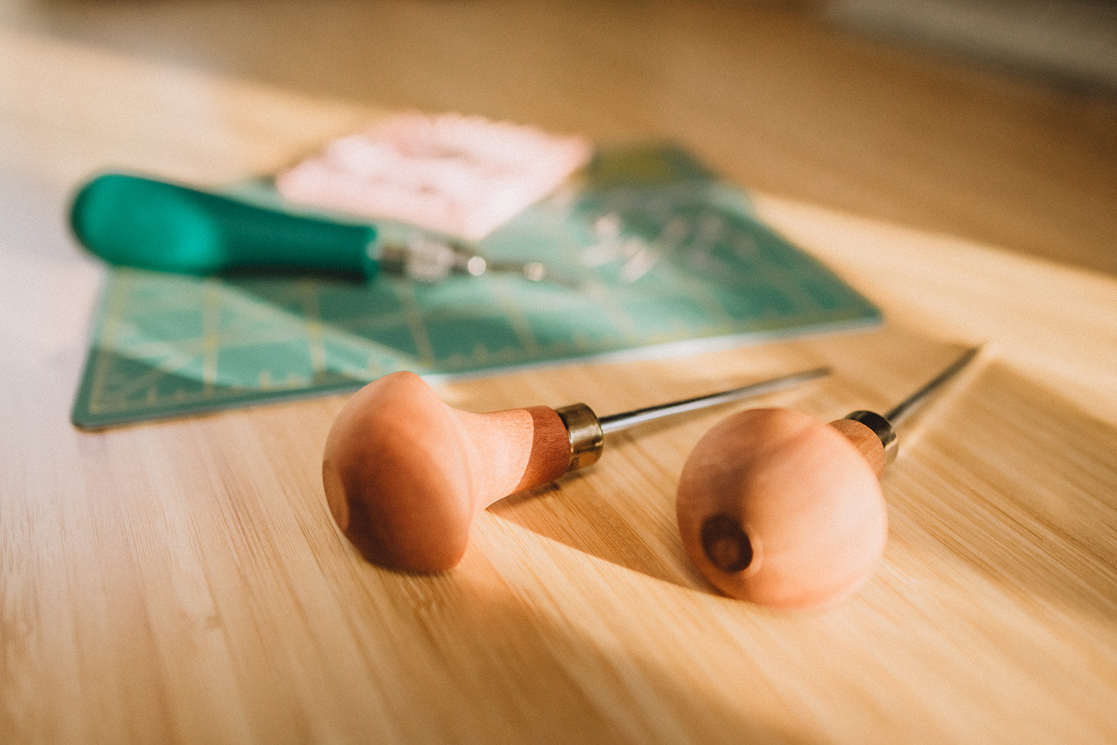 Two wooden-handled leatherworking tools on a wooden surface with a green leather square in the background.