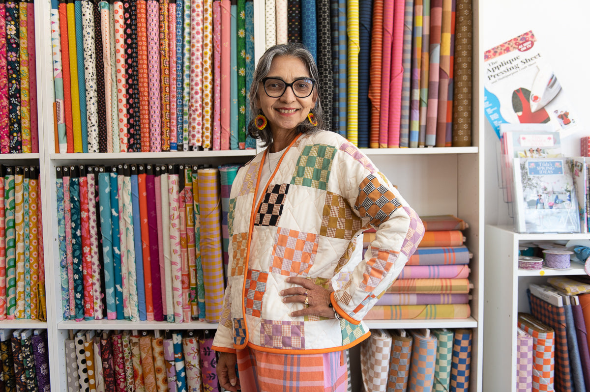 Woman wearing a checkered quilt coat standing in front of a colorful array of fabric swatches on shelves.