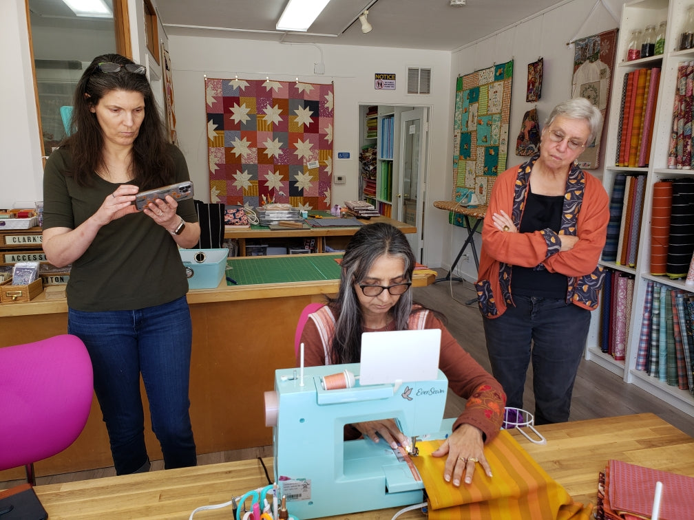 2 people watch a woman working at a sewing machine