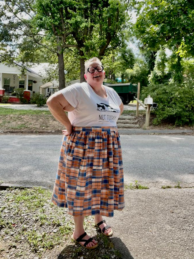 Person wearing a plaid skirt and graphic t-shirt standing on a street with trees and houses in the background.