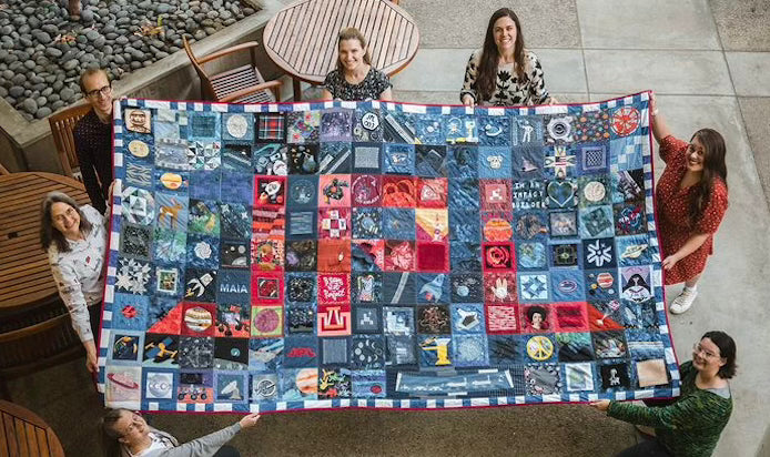 Group of people holding a large quilt with various patterns and colors.