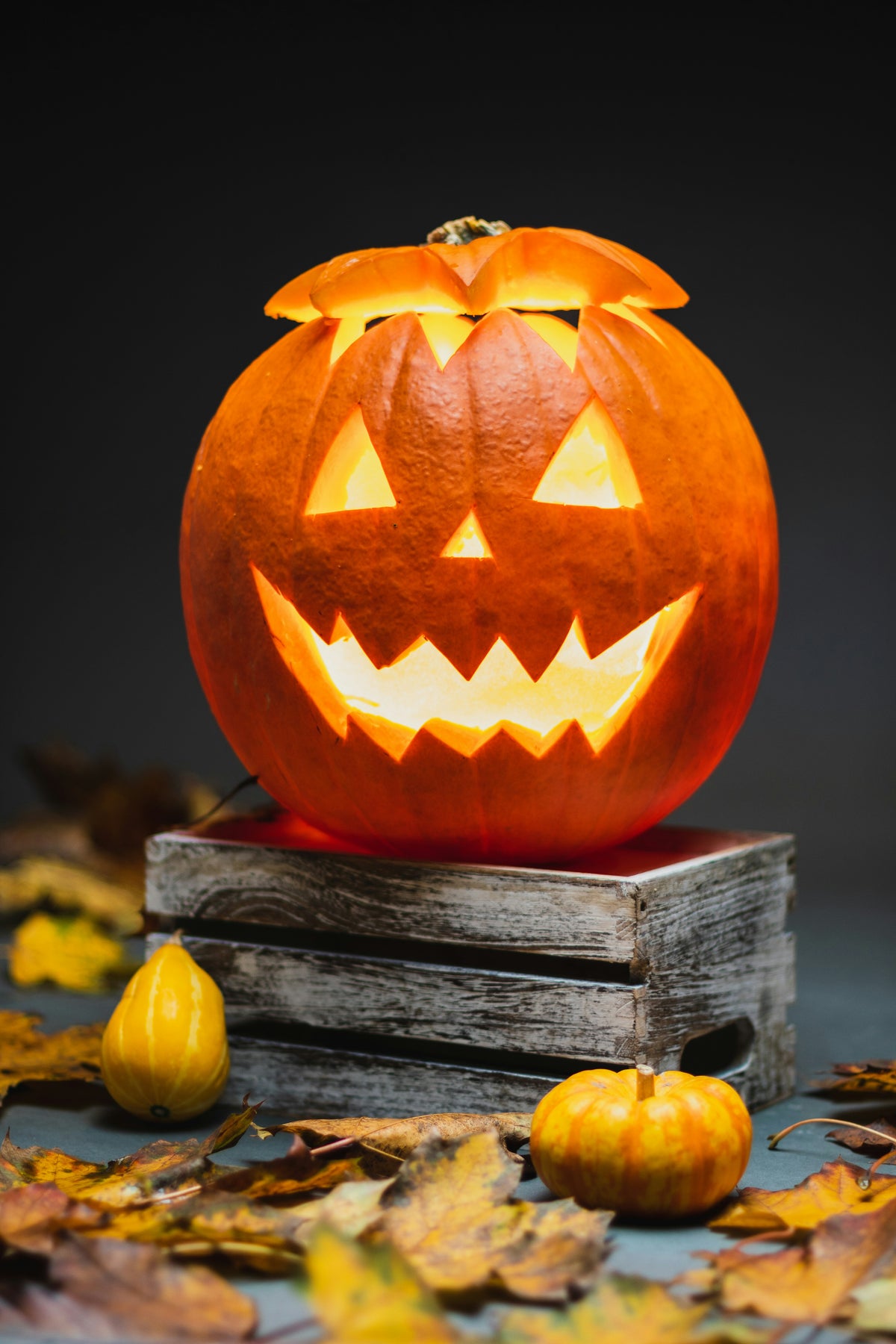 Carved pumpkin with a jack-o'-lantern face on a wooden crate surrounded by autumn leaves.