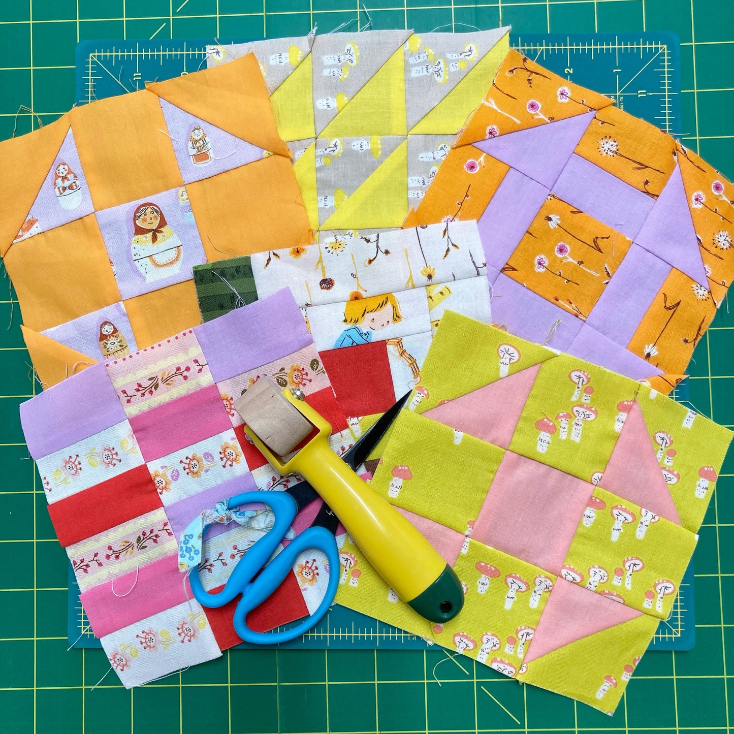 Various hand pieced quilt blocks on a cutting mat with scissors and seam roller